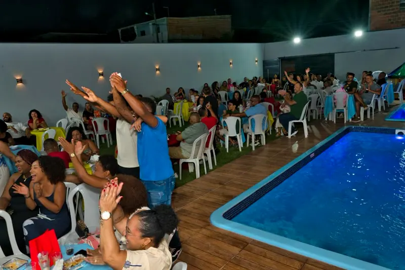 Festa à noite, com convidados nas mesas do gramado, ao lado da piscina, e as mãos para o alto