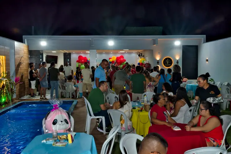 Festa de aniversário à noite, com a área coberta cheia de gente, balões e a piscina à esquerda