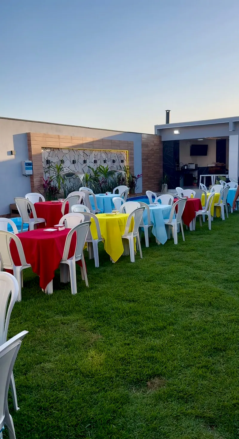 Mesas com toalhas vermelhas, azuis e amarelas no gramado, com a parede de pedra e a piscina ao fundo